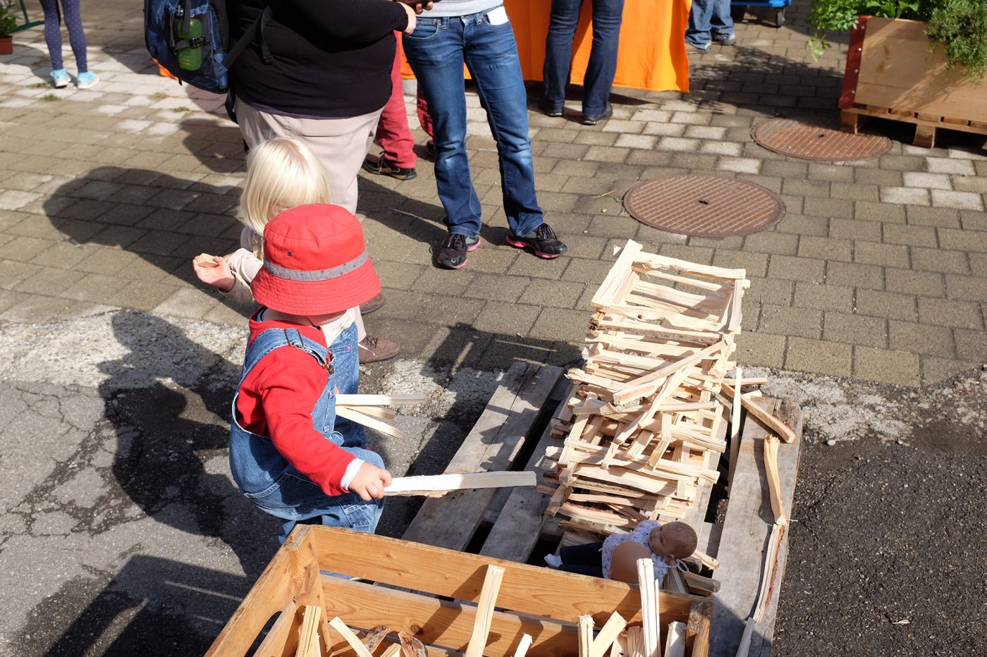 Ein kleines Kind mit rotem Sonnenhut hebt kleine Holzstückchen auf das Fundemant eines entstehenden Holzturmes.